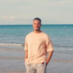 pexels-photo-33944932-33944932 A young adult male stands happily on a serene beach with clear blue water.